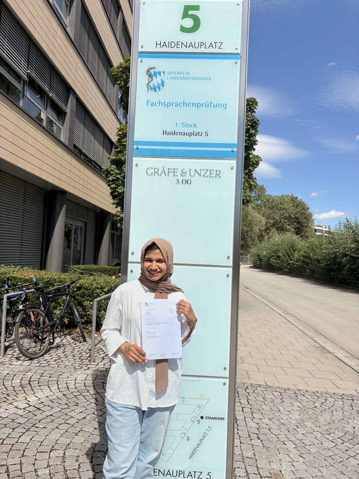 Nafeesa holding a document beside the Fachsprachenprüfung sign at the Bavarian Medical Association